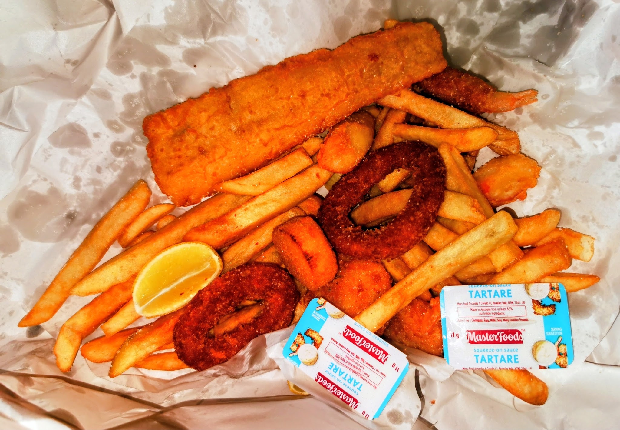Battered fish, calamari rings, chips, lemon and tartare sauce on white paper
